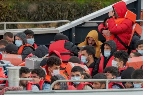 مجموعة من المهاجرين يرتدون سترات النجاة، يجلسون في قارب أثناء عبورهم القناة الإنجليزية، وسط أجواء من القلق والتوتر.