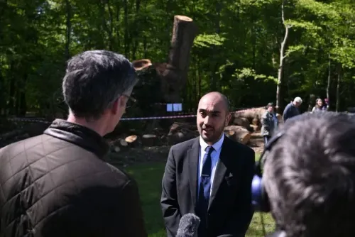 رجل يتحدث أمام الصحفيين حول قطع شجرة بلوط قديمة في لندن، مع بقايا الشجرة خلفه، مما يبرز تأثير هذا الحدث على المجتمع.