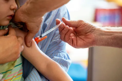 طفل يتلقى لقاحًا، حيث تُظهر الصورة يد ممرضة تحمل حقنة وتوجهها نحو ذراع الطفل، مع وجود شخص بالغ يساعده.