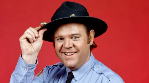الممثل ريك هيرست، المعروف بدوره في "The Dukes of Hazzard"، مبتسم ويرتدي قبعة شرطي، على خلفية حمراء.