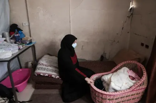 امرأة ترتدي كمامة تجلس على الأرض في شقة نصف مهدّمة، تحمل مولودها الجديد بجانب سرير صغير. تعكس الصورة معاناة الأمهات في غزة بسبب الظروف الصعبة.