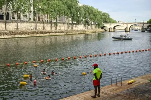 سباحون يستمتعون بمياه نهر السين في باريس تحت إشراف منقذ، مع وجود حواجز أمان ومشهد تاريخي للمدينة.