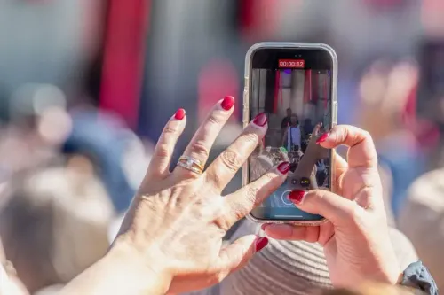 يد تحمل هاتفًا ذكيًا لتسجيل فيديو لمغني على المسرح، مع التركيز على تفاصيل اليد والأظافر المطلية باللون الأحمر.
