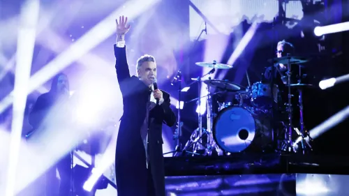 روبي ويليامز يؤدي على المسرح، مع إضاءة ساطعة وأعضاء فرقة خلفه، حيث يتحدث عن تحديات صحته النفسية ونقص فيتامين C.