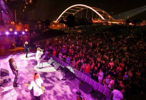 حفل موسيقي في الهواء الطلق يضم فرقة تؤدي على المسرح أمام حشد كبير، مع إضاءة ملونة وجسر في الخلفية.