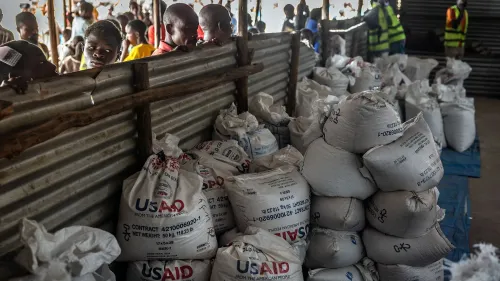 تظهر الصورة مجموعة من أكياس المساعدات التابعة للوكالة الأمريكية للتنمية الدولية (USAID) في مركز توزيع، مع وجود أشخاص في الخلفية ينتظرون الحصول على المساعدات.