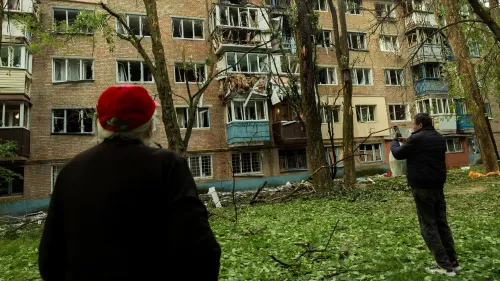 تظهر الصورة شخصين يقفان في حديقة أمام مبنى سكني متضرر، حيث تضررت النوافذ والأجزاء الخارجية بسبب الهجمات الروسية على كييف.