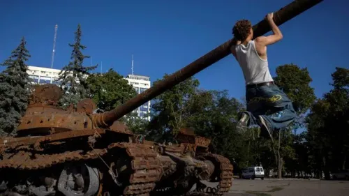 شاب يتدلى من مدفع دبابة قديمة في ساحة عامة، تعكس الصورة آثار الحرب والتوترات الجارية في أوكرانيا.