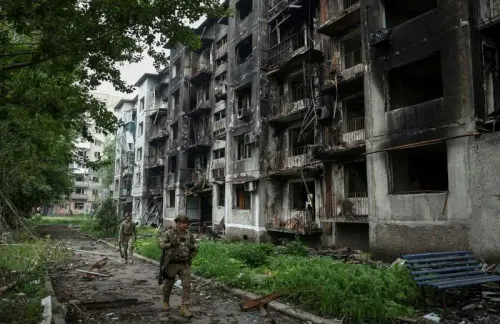 جنود يسيرون بجانب مبانٍ سكنية مدمرة بفعل الحرب، مع آثار الدمار والنباتات الخضراء المحيطة، تعكس الوضع الأمني المتوتر على الحدود الروسية الأوكرانية.