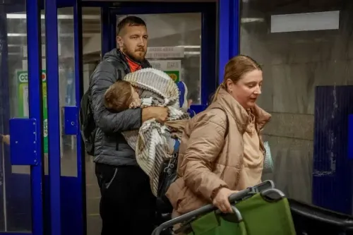 رجل يحمل طفلاً مغطى ببطانية، بينما تخرج امرأة مع عربة تسوق، في مشهد يعكس تأثير الحرب على المدنيين في أوكرانيا.