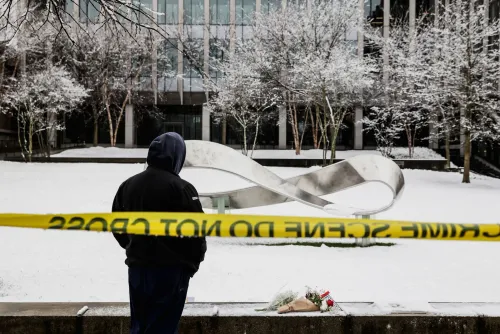 شخص يقف أمام موقع الحادث في جامعة براون، محاطًا بشريط الشرطة، مع زهور موضوعة على الثلج، مما يعكس القلق بعد إطلاق النار.