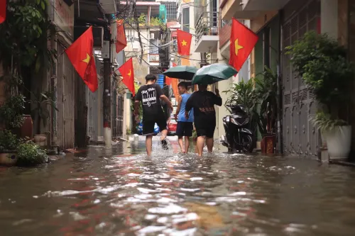 شارع مغمور بالمياه بسبب الفيضانات، حيث يسير ثلاثة أشخاص تحت المظلات، مع وجود أعلام حمراء تزين المباني المحيطة.