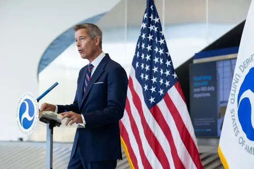 وزير النقل شون دافي يتحدث أمام ميكروفون في مؤتمر صحفي، مع العلم الأمريكي وعلم وزارة النقل خلفه، وسط أجواء رسمية.