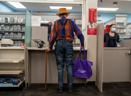 رجل مسن يقف في صيدلية، يرتدي قبعة وقميص ملون، ممسكًا بعصا وحقيبة تحمل شعار NYU Langone Health، بينما يتحدث مع موظفة.