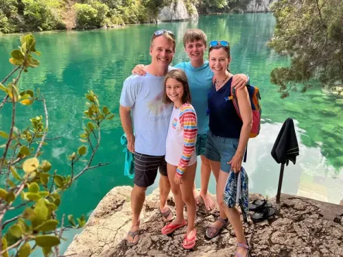 عائلة ألموند تتجمع بجانب بحيرة زرقاء، حيث يظهر الوالدان وأطفالهما في لحظة سعادة خلال مغامرتهم التعليمية حول العالم.