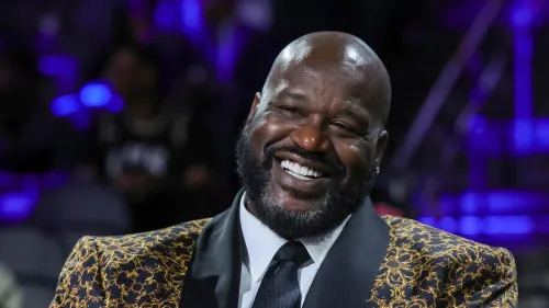 شاكيل أونيل مبتسم خلال حدث رياضي، يرتدي بدلة أنيقة، بينما يجلس في أجواء احتفالية مع إضاءة ملونة، مما يعكس روح الدعابة في برنامجه.