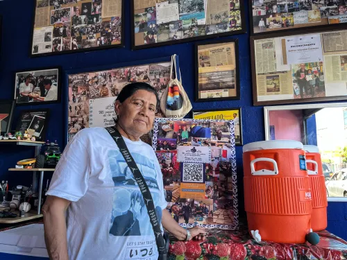 إستر موراليس تقف في مطعمها بتيخوانا، محاطة بصور ومقالات تبرز إنجازاتها في دعم المهاجرين من خلال مشروعها "طعام دافئ".