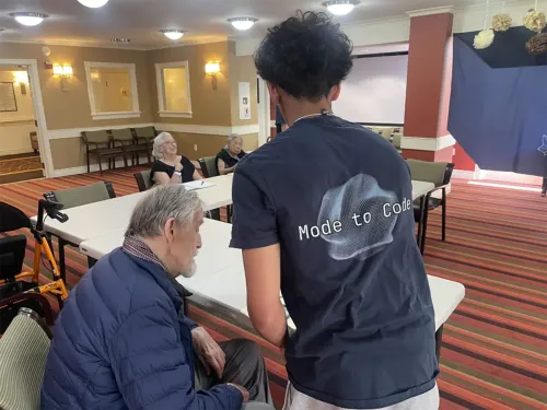 شاؤول، طالب في السابعة عشرة، يساعد كبار السن في تعلم التكنولوجيا خلال دورة "مود تو كود" في مركز للمسنين، حيث يظهر تفاعلهم الإيجابي.