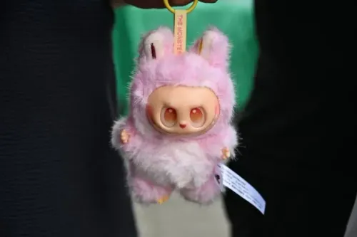 دمية صغيرة ذات شكل مميز ولون وردي، ترتدي زيًا يشبه حيوانًا، تحمل علامة "THE MONSTER"، تُظهر الابتكار في تصميم الألعاب.