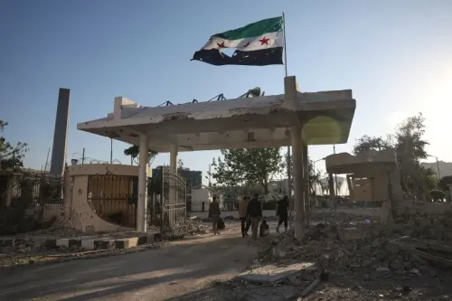 مدخل مدمّر لموقع عسكري في سوريا، يرفرف فوقه علم الثورة السورية، مع أشخاص يسيرون في الخلفية.