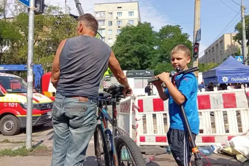 رجل وطفل يقفان بجوار دراجتين في موقع تعرض لهجوم، مع وجود سيارات إسعاف وخدمات الطوارئ في الخلفية، يعكسان آثار الحرب في أوكرانيا.