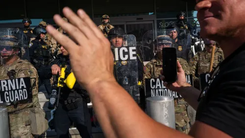 مشهد من احتجاج في لوس أنجلوس يظهر أفراد الحرس الوطني والشرطة، مع شخص يحمل هاتفه لتوثيق الأحداث.