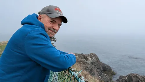 رجل يرتدي سترة زرقاء وقبعة، يستند إلى سياج قرب ساحل ضبابي، مما يعكس الهدوء والطبيعة المحيطة به.