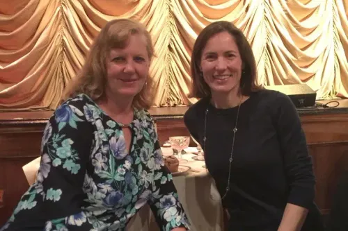 صورة لامرأتين تجلسان معًا في مكان مغلق، مع خلفية من الستائر، تعكس أجواء اجتماعية.