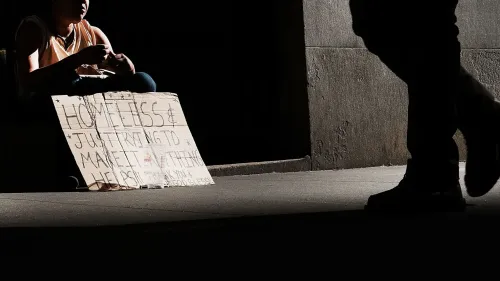 شخص بلا مأوى يجلس في الظل يحمل لافتة يطلب المساعدة، تعكس واقع التفاوت الاقتصادي الحاد في الولايات المتحدة.