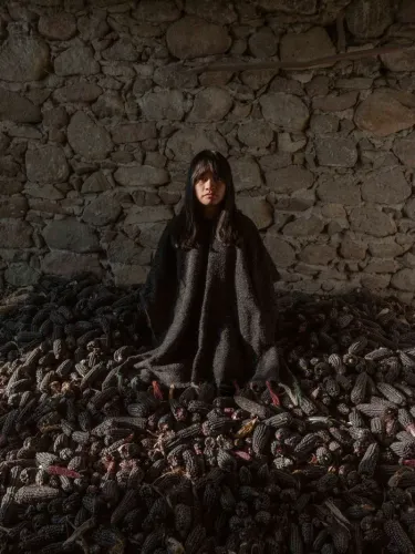 امرأة تجلس وسط كومة من حبوب الذرة السوداء في مكان ذو جدران حجرية، تعكس صورة من الحياة التقليدية والتراث الثقافي.