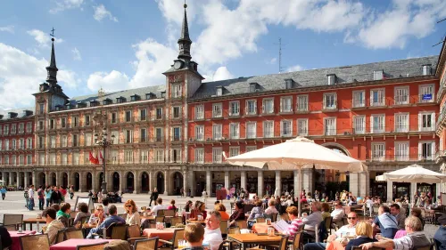 مجموعة من الزوار يستمتعون بالغداء في مطعم خارجي في ساحة بلازا مايور بمدريد، تظهر العمارة الأنيقة والسماء الزرقاء.