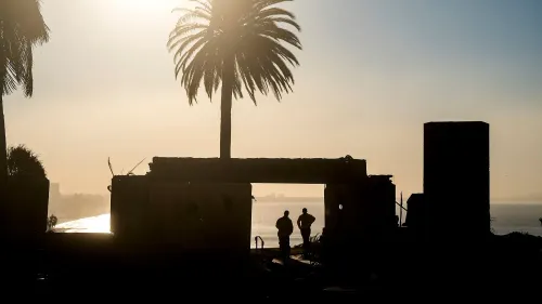 دخان يتصاعد من خلف أنقاض منزل محترق، بينما يقف شخصان في الظل، مما يعكس تأثير حرائق الغابات في لوس أنجلوس.