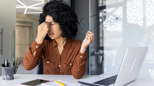 امرأة تعاني من الإجهاد أثناء العمل، تمسك نظاراتها وتضع يدها على جبهتها، محاطة بمكتب يحتوي على حاسوب محمول وأدوات مكتبية.
