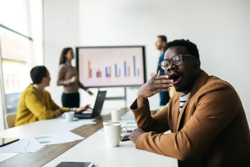 رجل في اجتماع يبدو متعبًا ويشعر بالنعاس، بينما يعرض شخص آخر بيانات على شاشة كبيرة.