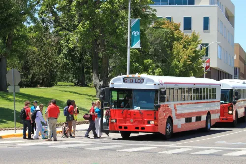 مجموعة من الطلاب ينتظرون حافلة في حرم جامعة نيو مكسيكو، مع وجود مباني خضراء وأشجار في الخلفية، في إطار نشاطات أسبوع التوجيه.