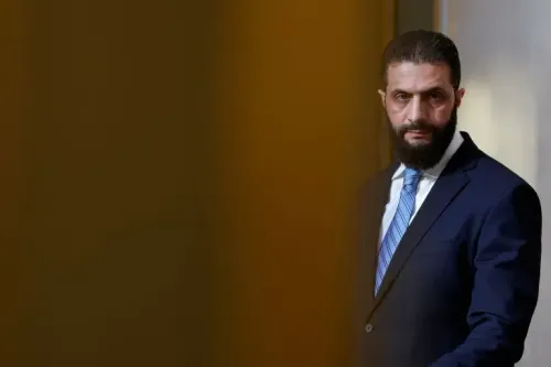 رجل يرتدي بدلة رسمية، يبدو جادًا، يقف في مكان مغلق، مع خلفية غير واضحة، مرتبط بالانتخابات البرلمانية في سوريا.