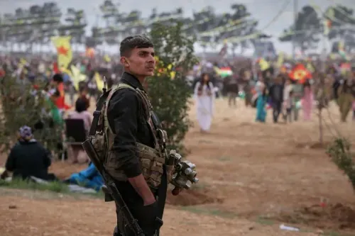 جندي من قوات سوريا الديمقراطية يرتدي زيًا عسكريًا، يقف في مواجهة تجمع حاشد في محافظة حلب، وسط توترات متزايدة مع القوات الحكومية.