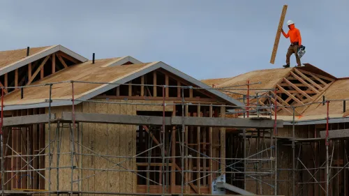 عامل بناء يرتدي سترة برتقالية وقبعة، يعمل على سقف منزل قيد الإنشاء، مع وجود سقالات وألواح خشبية حوله، مما يعكس النشاط في سوق العمل الأمريكي.