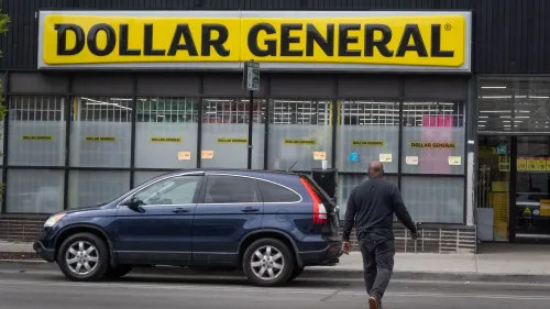 واجهة متجر "دولار جنرال" مع شعارها البارز، يظهر رجل يسير نحو المدخل، مما يعكس زيادة الإقبال بسبب الظروف الاقتصادية.