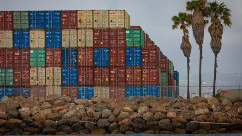 حاويات شحن ملونة متكدسة في ميناء أمريكي، مع صخور على الواجهة البحرية ونخيل في الخلفية، تعكس حركة البضائع المتأثرة بالتعريفات الجمركية.
