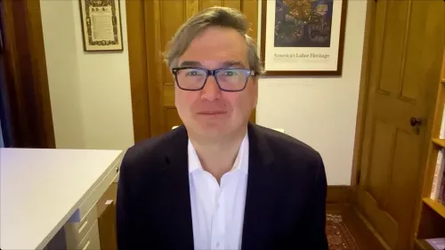 رجل يرتدي نظارات ويبتسم، يجلس في مكتب مع خلفية تحتوي على لوحات وصور تتعلق بالتراث الأمريكي.