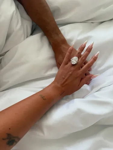 خاتم خطوبة كبير ومبهر مرصع بألماس، يُظهر يد امرأة تتشابك مع يد رجل، مع خلفية من الأغطية البيضاء.