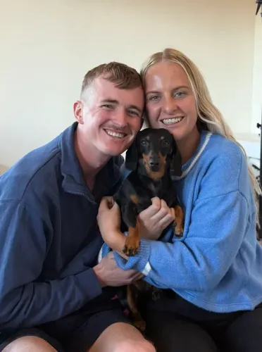 فاليري، الكلبة الألمانية، محاطة بمالكيها جورجيا وجوش، تعبيرات السعادة تظهر على وجوههم بعد لم الشمل.