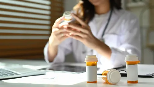 امرأة ترتدي معطف طبي تفحص زجاجة دواء على مكتب، مع زجاجات دواء أخرى في المقدمة، تعكس موضوع الرعاية الصحية عن بُعد.