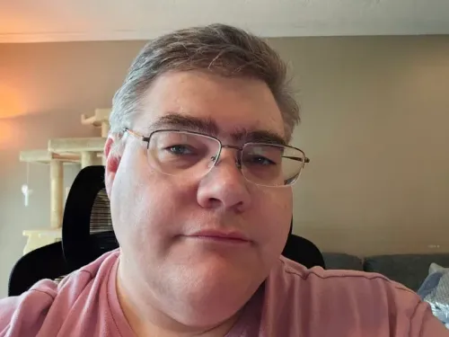 صورة لرجل في منتصف العمر يرتدي نظارات، يجلس في غرفة مع أثاث بسيط، يعبر عن قلقه بشأن ارتفاع أسعار التسوق عبر الإنترنت.