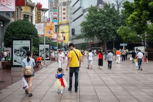 رجل وطفلة صغيرة يسيران في شارع مزدحم في بكين، مع لافتات ملونة ومتاجر في الخلفية، مما يعكس الحياة الحضرية والتحديات السكانية.