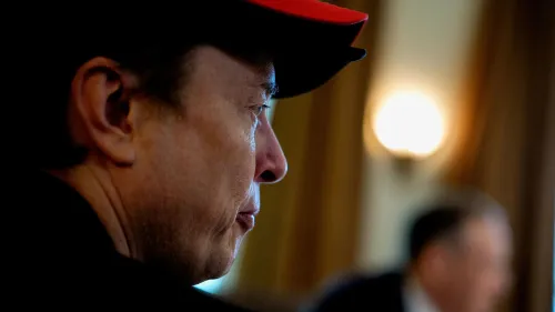 إيلون ماسك، الرئيس التنفيذي لشركة تسلا، يظهر من الجانب أثناء اجتماع، مع التركيز على تعبير وجهه وارتداءه قبعة حمراء.