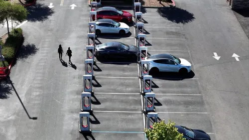 تظهر الصورة موقف سيارات يحتوي على عدد من سيارات تسلا في صفوف منظمة، مع وجود شاحنتين كهربائيتين في الخلف.