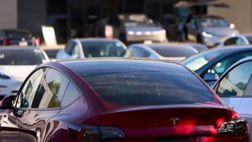 سيارة تسلا حمراء متوقفة في ساحة عرض، مع سيارات أخرى في الخلفية، تعكس تفوق تسلا في تصنيع السيارات الأمريكية.