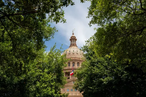 مبنى الكابيتول في تكساس محاط بأشجار خضراء، مع العلم الأمريكي وعلم تكساس، مما يعكس الأحداث السياسية الجارية حول إعادة تقسيم الدوائر الانتخابية.